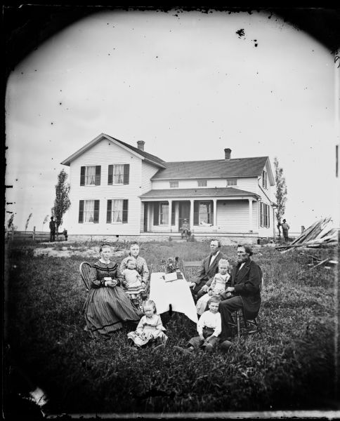 A family is seated around a table upon which are books and flowers. Behind them is a frame house with a person sitting on its porch.