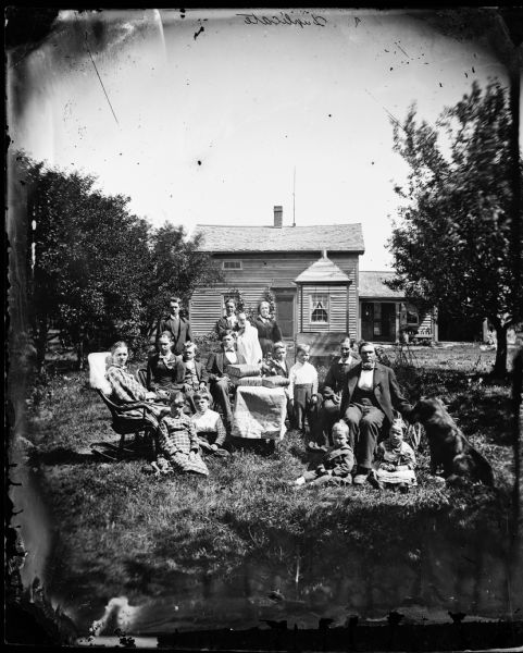 A large family group and a dog are posed around a table that has large books on it. The frame house behind them has a sway in its roof. A well with a wood enclosure and a roof is in the background near the front door. Above the front door is a Hekla Fire Insurance sign.
