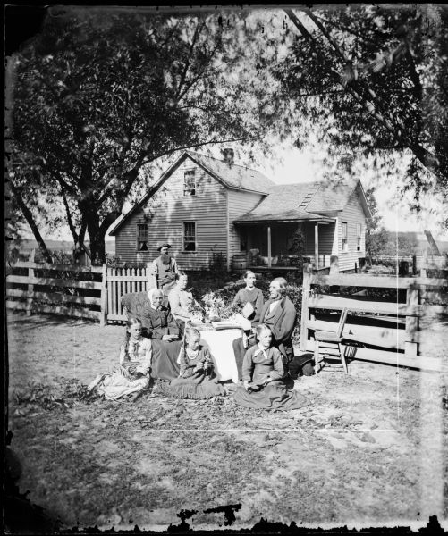 A family of seven is sitting in the foreground around a table covered with books and flowers. Three young girls are sitting on the ground in front of the table, and an older man, holding a chisel hammer, and a woman holding a book are sitting on either side of the table behind the girls. Two women, one holding a hand fan, are sitting behind the table, and a man with a long beard and wearing a hat is standing near the gate holding a ball peen hammer. Behind the group is a shady yard with a picket fence and a frame house with a ladder laying flat on the roof behind the porch roof. A barrel is near the corner of the house near the porch.