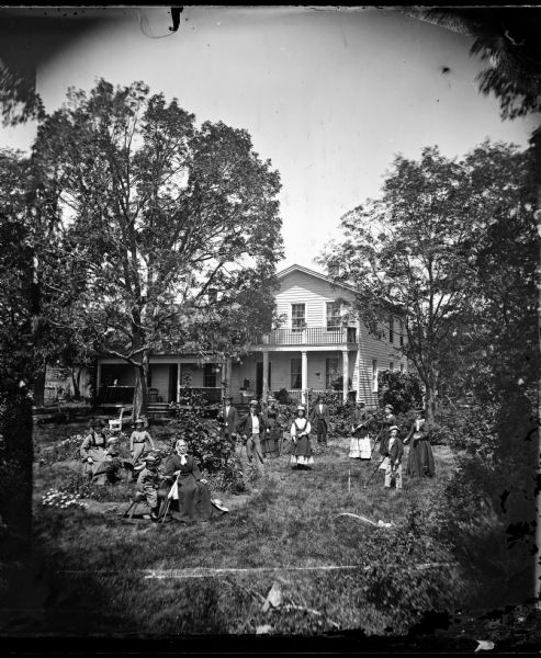 Herman Amberg Preus, his wife Caroline Keyser Preus, and their family, are in the yard playing croquet. One woman sits with a closed parasol in her hand, and next to her is a boy on a rocking horse. The frame house behind them has latticework on its porch and a fence style balcony on the porch roof.