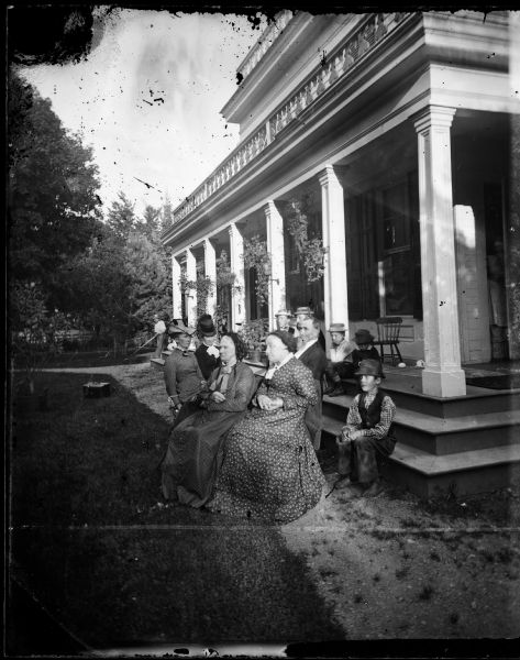The Edwin and Angeline Coe Hillyer family is grouped at the corner of a large Greek Revival house with ornamental railing work on its porch roof. A man is using some sort of yard tool in the left background.  Angeline is on the far left. Edwin has a beard and sits near the steps.