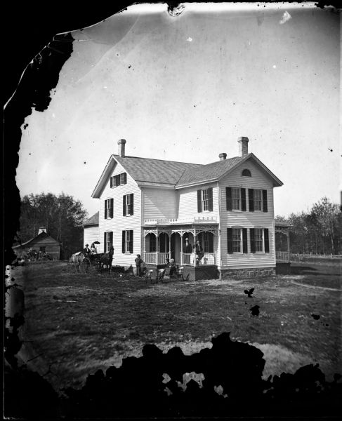 A family of five is posed around a large frame house with carpenter's lace on its porch, a fan window in the gable end, half windows on the attic story on one side and a second porch in back with carpenter's lace and latticework. A man and woman sit in a carriage next to the house. A shed and woodpile are in the background on the left.