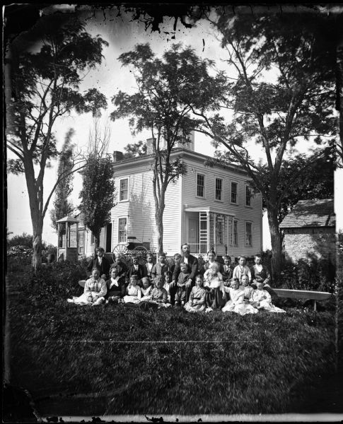 Twenty-one children are seated on wooden benches and the ground in front of a square frame house with a hip roof and cupola. The Reverend Erik Jensen (1841-1927) and his wife Petronelle Brevig Jensen sit among the children. In front is a small toy wheelbarrow. Behind them is a carriage, a well, a barrel and on the right a small stone outbuilding. 
Jensen wrote song books and was the pastor at Mound Springs from 1870-73, at Jefferson Prairie in Rock County from 1874-82 and at Coon Valley from 1882-93. He made a trip back to Norway in 1873-74, having immigrated in 1867.