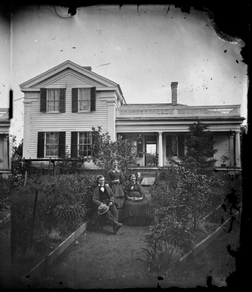 Two women and a man posed in the garden path of an L-shaped Greek Revival frame house. The front and back doors are open, revealing a breezeway straight through the house. This is the G. Bashford home, now on highway 19, in section 7, located in "Madena".