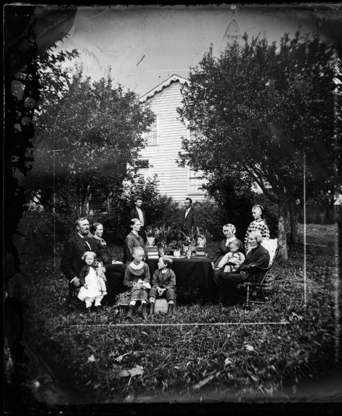 A family is gathered outdoors around a table with plants and books. A girl holds a doll and next to her a boy sits on a soap box. The frame house behind them is obscured by trees.