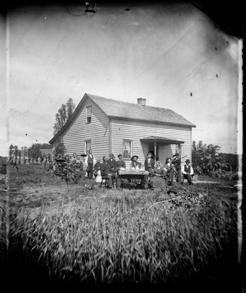 A family is gathered around a table with dishes on top. To the right is a man sitting and holding a violin, and to his right a boy is holding a gun. The house behind them has a salt-box roofline, and a roof-covered entrance. In the foreground on the right is a man wearing a hat is lying in the bushes holding a switch.