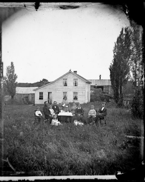 A family is seated around a table while a woman pours the coffee. Behind them, two people stand in the doorway of a frame house. A man stands on the roof of a farm building in the background on the right, and another man stands on hay piled up in a wagon.