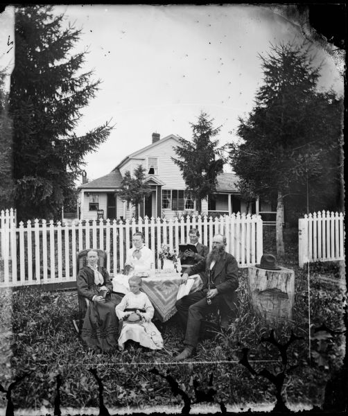 A family is sitting around a table in front of a picket fence. Three of the adults are holding books, one man is holding a long pipe and a girl is holding her hat in her lap. To the right is a hat resting on a mounting block (tree stump). The frame house behind them has latticework on the sides and top of a small porch and on the bottom of a larger porch.