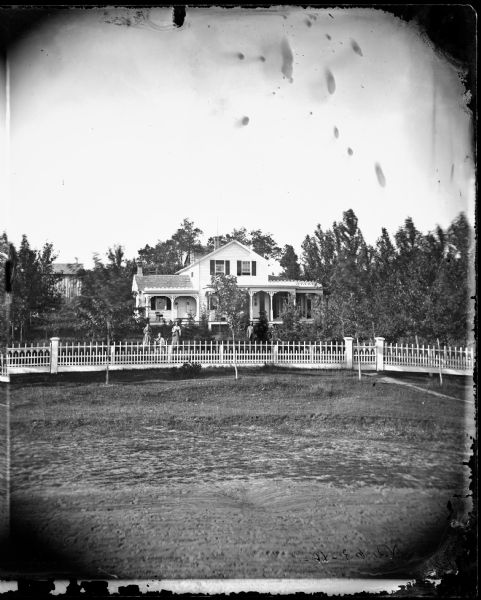 The Samuel Barber residence is a frame house with carpenter's lace, porch roof trim, latticework at the bottom of the porch and a picket fence in the foreground. A group of people are standing behind the fence.