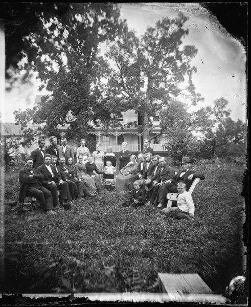 Herman Amberg Preus, his wife Caroline Keyser Preus and their visitors are seated in a large semicircle with baby in carriage in center; frame house with carpenter's lace and railing on front porch behind. This is the second parsonage of Pastor Herman Amberg Preus. Text on back of PH 4252 print reads: "Nels Dahl 3rd from left standing, Ingeborg Dahl seated with hand on carriage, Louis Dahl - seated on ground right, John J. Anderson 2nd from right seated, H. A. Preus 5th fro right seated, Agnes W. Preus to left behind carriage".