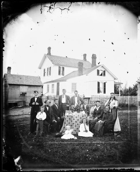 A family is gathered around a table in the yard. Behind them is a fence and behind that a frame house is seen in a three-quarter view. It has arched second story windows with shutters on the side, and a small frame building behind it. A small building is at the end of the fence on the left.