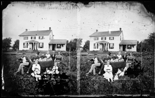 A family is grouped around a table, including a man with a top hat, two dolls on the ground between two small girls and a woman holding a closed fan. Behind them on the left is a pump. The frame house has a bird cage on the porch and half windows on the small second story.