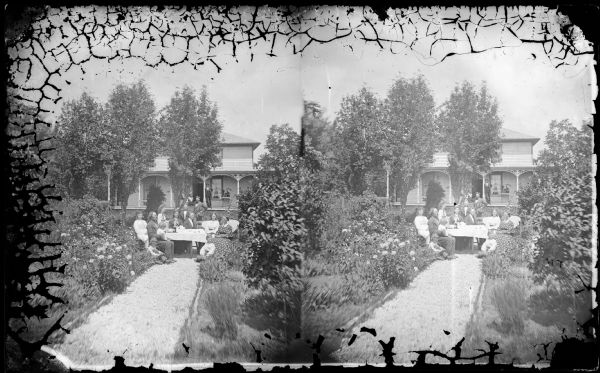 A family is seated around a table in the middle of a garden path, and a man stands on the porch of the frame house behind them. The house has carpenter's lace and latticework at its foundation.
