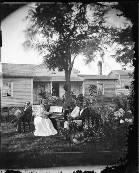Two men, one of whom is Halver Skavlem, and a woman, Gunil Skavlem, are posed in a yard. One man reads the "Western Rural", a newspaper dated August 14, 1875. The woman plays the melodeon and the other man holds his hat and a newspaper. A broad-rimmed hat is in the foreground bushes, and another hat is hanging from the tree trunk in the center. Behind them is a single story frame house with vines on its porch. Another person is sitting on the porch.