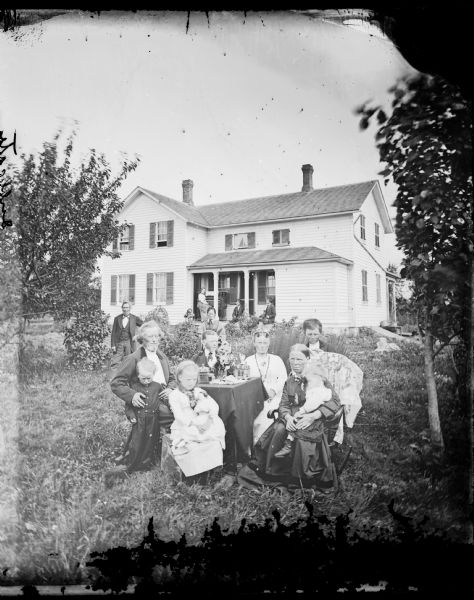 A family is in the yard around a table. A girl holds her doll in her lap. Behind them are two people standing in the yard, and more people on the porch of the house on which a piano is sitting.