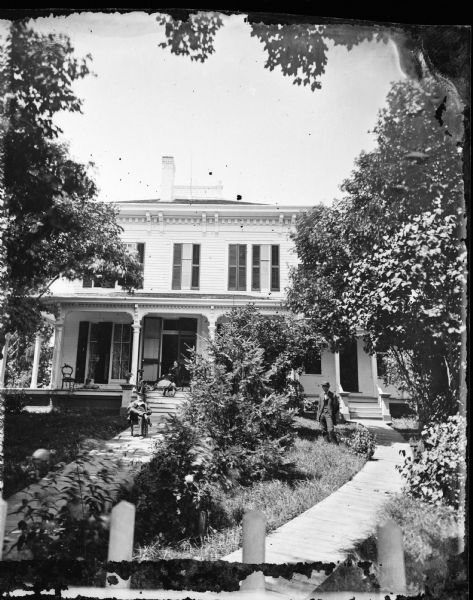 Men stand in the yard, and a woman stands on the porch of a frame house with double brackets, double front doors with screen doors and a double walkway leading to it.