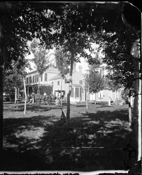 A family stands around a carriage in the yard of a two-story frame house with bay window and multiple additions. Vines grow on the side of the front porch and there is a bell on the roof of the house. In the foreground a turkey stands near a tree.