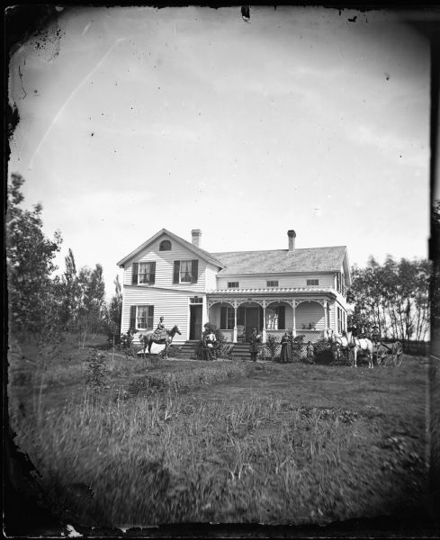 On the left a woman sits side saddle on a horse, and on the right two men are on a wagon hitched to two horses. Between them are a group of men in front of an upright and wing house with a porch heavily decorated with latticework and a small parapet on its roof. In addition, the house has a fan shaped window in its attic story.