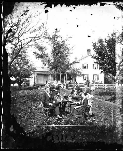 A family is in a yard around a table with books on it. One woman holds a fan, another sits in a rocking chair with a shawl on its back. A fence and open gate are behind them. The frame house in the distance has cutwork porch posts.