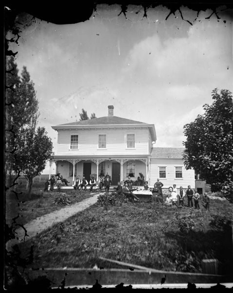 An extended family of twenty-one people is gathered in the yard and on the porch of a large bracket style frame house that has carpenter's lace on its porch, latticework at bottom of the porch, a stone foundation and a frame addition on the right. Leading to the house is a wooden walkway.