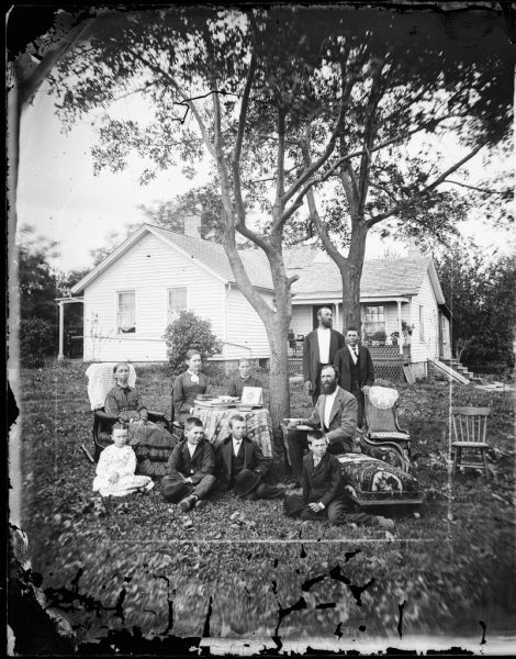 A family is posed around two trees in a yard. A man sits on a chaise lounge and a woman in a striped dress sits in a rocking chair with a shawl on its back. The frame house behind them has a sewing machine on its porch and latticework in evidence. A bulkhead basement entrance is on the right side of the house near steps that lead up to a door.