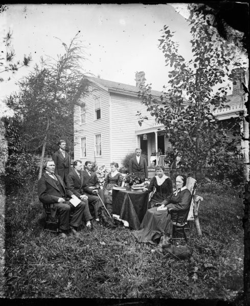 The R. Daniel Rickelsen Rickelsen family is gathered around a table in a yard. A man holds a stick, and a clay pipe sits on the table. The frame house behind has a hanging bird cage. Its porch features a decorative railing and trim along the edge of the porch roof.

