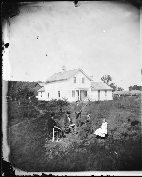 Two women and three men are posing in a yard. One man is holding a shotgun, another a carpenter's saw. The frame house behind them has a small front porch with a railing on both sides. A large haystack is on the right behind the house, and a barn is in the background on the left.