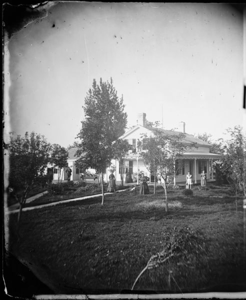 Four women are posing in a yard among trees, and one woman is sitting on the porch of the house. A dog is sitting in a chair in the yard near one of the woman. There is a man on the left corner of the house standing and holding a shovel. Two children are standing on the porch roof of the frame house, which has several additions. The porch is continuous along two sides of the house.