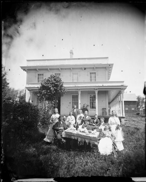 A large family group is gathered around a table for coffee. The women are holding bunches of flowers, and behind them is a large bracket style house with a porch on at least two sides. A farm building is in the far background on the right.