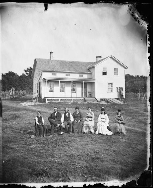 A young boy, two men and four women are seated in a row the yard of a frame house with latticework on the porch foundations, brick steps on the side, small windows in front on the second-story and a visible entrance to the basement in front.