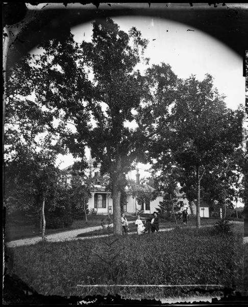 A man with a beard is sitting in a chair next to a boy and a girl, all posing with an informality unusual for a Dahl photograph. Behind them a younger man is carrying a smaller child toward the foreground group. A large house, obscured by large trees, is further back.