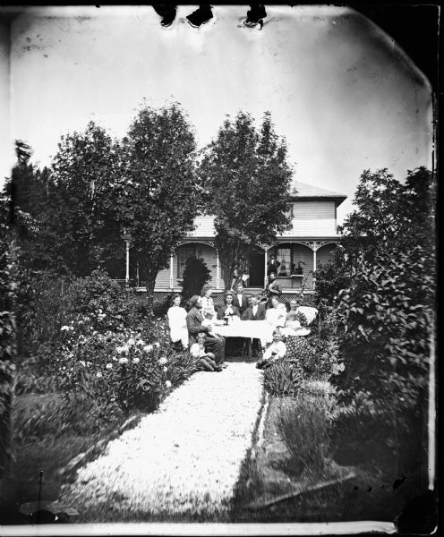 A family sitting around a table in a garden path. A man is standing in the background on the porch of the frame house. The porch has carpenter's lace above, and latticework at the bottom.