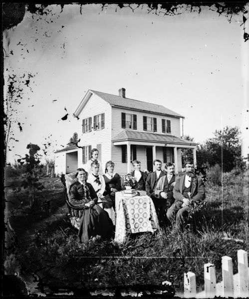 A picket fence is in the right corner of the foreground. Behind it is a family gathered formally around a table in the yard of a frame house with a shallow porch.
