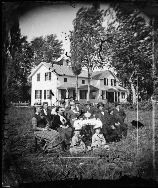 A large group of people sitting around a table in the yard. The men and women are wearing hats. The table has a large piece of ornamental glassware on it. The large frame house behind them has a sofa on its front porch and a framed picture. There are small shuttered windows above the porch and a small side porch with latticework.