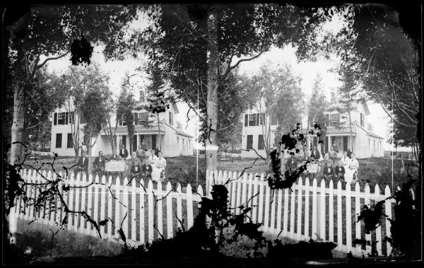 Family behind picket fence sitting around a table. Behind them is a man leaning on tree and one man standing on the porch of a frame house. The house has nice cut work above the front door and carved work on the door. Some of the shutters on the windows are closed. There is a small one story addition behind the house. Clothes are hanging on a line in the right background.