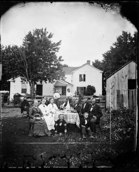 Family in yard around a table, with wood fence and frame house behind; one lady has a stereoscope in her lap. The house has an angular small addition with window and a shutter that is closed. There is a farm building just to the right side of the family.