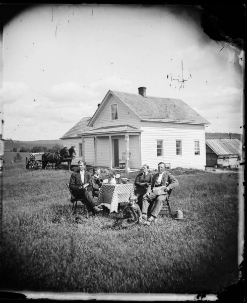 S. Pettersen family around square "center" table with a checkered table cloth in a house yard. A child is seated on the ground with the family dog while another family member holds a stereoscope. A mirror sits on the table. The upright-and-wing frame house has a rocking chair on the porch and the horse-drawn wagon is pulled up next to the house for inclusion in the picture.