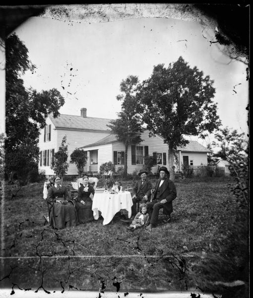 Family seated around a table in the yard of house. On the table are books, and a statue of a horse and rider.