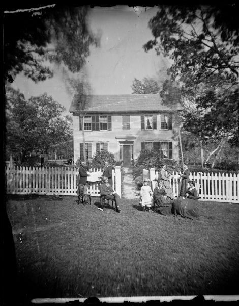 The Ole Wettlesen family poses in the yard in front of a picket fence with women knitting. Two standing men appear to be examining an unfolded map. The frame house behind has pedimented windows and glass trim at front door. Text on back of print of stereograph reads: "Son William Wettlesen holds plat book open to Dunirk township — William lived in Stoughton".