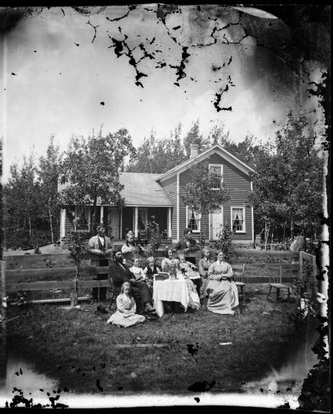 The Van Norman family seated outdoors in front of, and standing behind a fence with their frame L-shaped house behind.