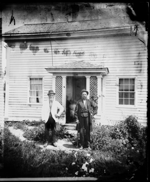Two men in hats, one with a walking cane, are standing in front of a frame house that has a small porch with latticework in front and a Hekla Fire Insurance sign above. Lars D. Reque is on the left.