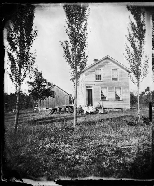 A couple with three children posing in a yard. The woman has a sewing machine, the man has a table saw and lathe. The two-story frame house has a glass panel above its front door, and a ladder is leaning against a tree. There is a frame outbuilding in the background. Lombardy poplars are in the foreground.