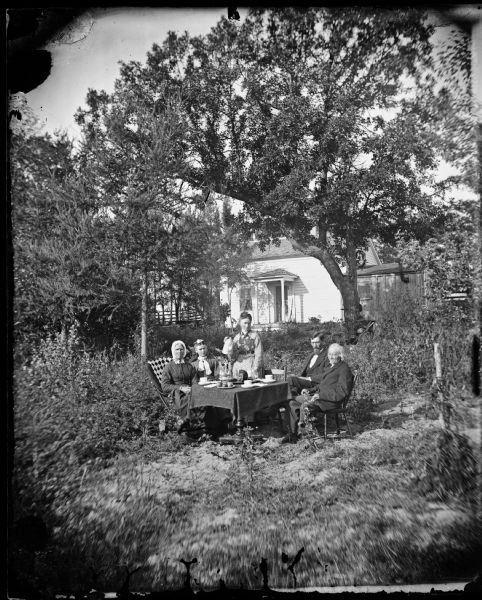 The Springer family sits in their yard around a table. A patterned shawl is on the back of a chair, and a girl has a scarf around her neck. A woman holds a coffee pot. The frame house behind is obscured by trees and has a small addition, possibly a kitchen that has a pipe chimney.