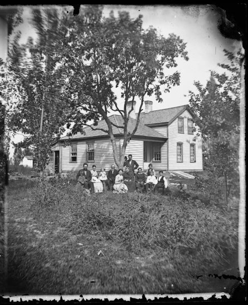 A family of twelve sitting in chairs under a tree in their yard. A child sits in a high chair. The frame house has a Hekla Insurance sign above its door; the windows in front have cut work above them and the basement entrance is visible.
