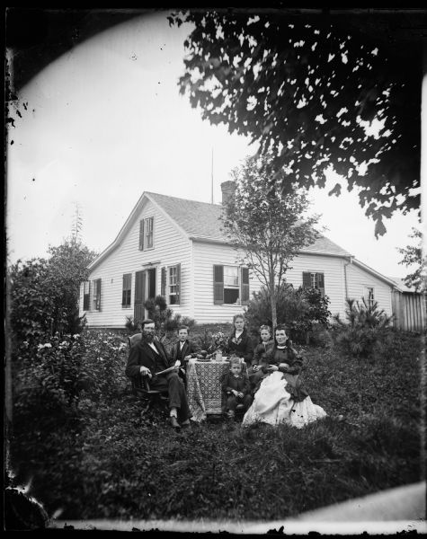 Two women, a young girl, two boys and a man are gathered around a table that has fruit displayed on plates. The man is sitting in an office chair. The frame house behind has a back-slanted roof and shutters for the windows and front door. A doll is propped up on the sill of an open window. An interesting tablecloth is on the table and small pine trees are nearby. An outbuilding is behind the house in the background on the right.
