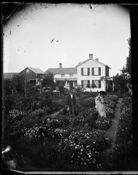 Elevated view of a couple standing in a garden. The woman holds an open parasol. The frame house behind has draped curtains and shades pulled on second story windows. There is a fence on the left side in front of the house and farm buildings in back.