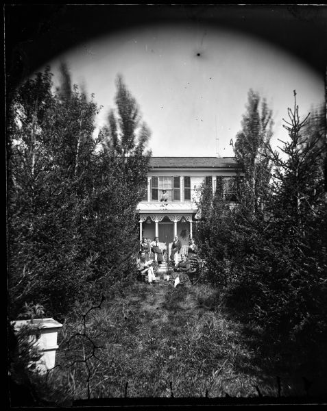 A family is sitting at the end of a tree-lined path on a hill leading up to a frame house with a porch. A boy is sitting on the porch roof, which is flat. He has two small animals in his lap. The group in the tall grass in front includes a woman sitting on the right with a parasol on a cushioned rocking chair, and a man with a long beard sitting on the left holding a book or newspaper. Two women are standing near the porch, and a man and a woman are sitting on the porch railing posts. Another woman standings near the porch on the right. Two birdcages are hanging on the porch. In the foreground on the left is a wooden box sitting on the ground which may be a beehive.