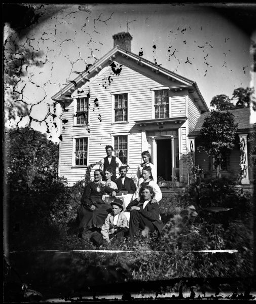 Riley, Springdale Township, Dane Co. Four men and four women around a table in yard in front of two-story Greek Revival frame house with a Madison Mutual sign above the door. Addison White (born in Vermont) settled the site in 1849. After he died (12/3/1873) his wife Sarah (born in New York) continued to farm the property. 