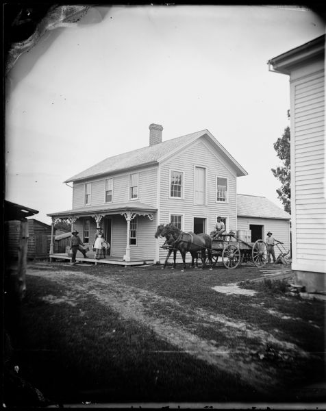 A farmer driving a horse-drawn wagon with milk cans. Behind him is a frame structure, possibly a cheese factory, with a porch and a full door on the second story. Another man is standing on the right behind the wagon. A man and woman are standing on the front porch of the building. Since this photograph was taken by Andreas Dahl, it is likely the scene was located somewhere in Dane County.