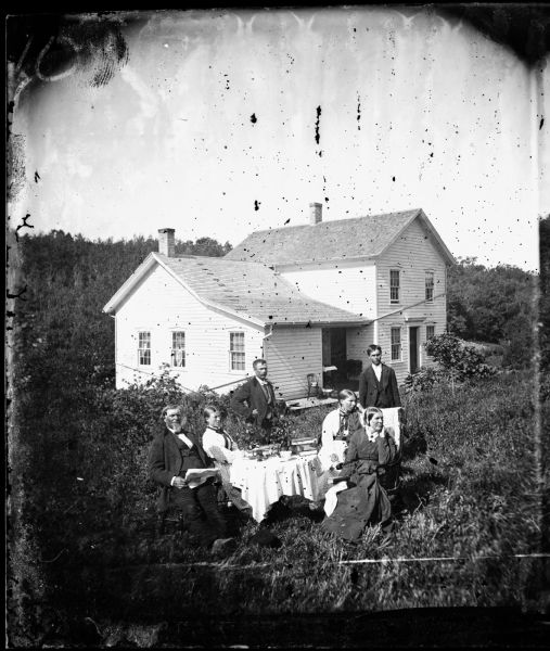 Left to right, standing: Hans A. Bringager and Erik Sviggum; Seated: C. Gulson, Annie Pedersen, Mary Gulson Brager and her mother Thora Gulson; all gathered around a table in the yard. A hat is on ground and the frame house stands with a chair, table and baskets on its porch.